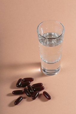 Diyet, beslenme, sağlıklı yeme konsepti. Bej arka plana kahverengi softgels kapsülleri dökmek için gıda takviyeleri ve bir bardak su dolu yağlı kapsüller..