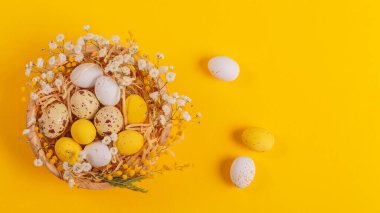 Easter candy chocolate eggs and almond sweets lying in a birds nest decorated with flowers and feathers on a yellow background. Happy Easter concept.