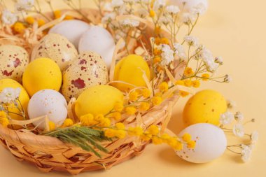 Easter candy chocolate eggs and almond sweets lying in a birds nest decorated with flowers and feathers on a yellow background. Close up. Happy Easter concept.