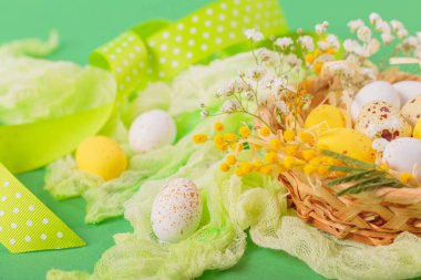 Easter candy chocolate eggs and almond sweets lying in a birds nest decorated with flowers and feathers on green background. Happy Easter concept.