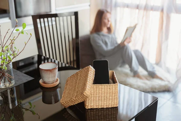 Smartphone is in separate wicker box on table. Woman reading a book in background. Stop using digital gadgets at home in favor of reading books and meditation. Mental and digital detox concept