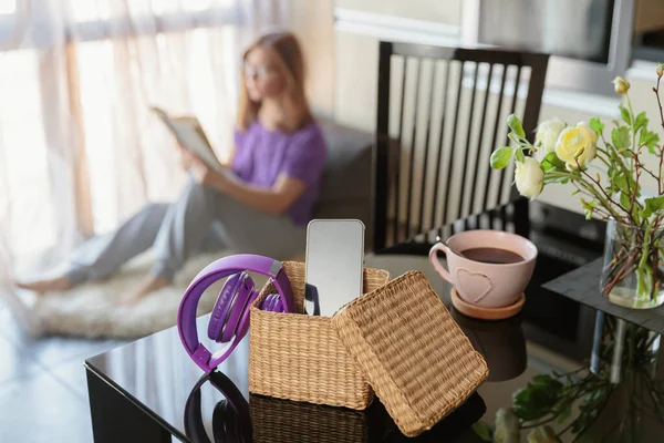 Smartphone is in separate wicker box on table. Woman reading a book in background. Stop using digital gadgets at home in favor of reading books and meditation. Mental and digital detox concept