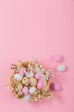 Easter candy chocolate eggs and almond sweets lying in a birds nest decorated with flowers and feathers on pink background. Happy Easter concept.