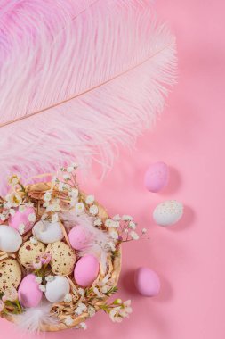 Easter candy chocolate eggs and almond sweets lying in a birds nest decorated with flowers and feathers on pink background. Happy Easter concept.