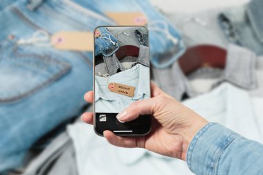 Kadın elinin kot kıyafetlerin fotoğrafını çekerek akıllı telefondan ikinci el moda uygulamasını yüklediği görüntüler. İnternette giysileri sat, al ve değiş tokuş et.