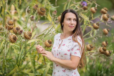 Young beautiful woman standing near big burdock plant and touching the leaves in old overgrown park