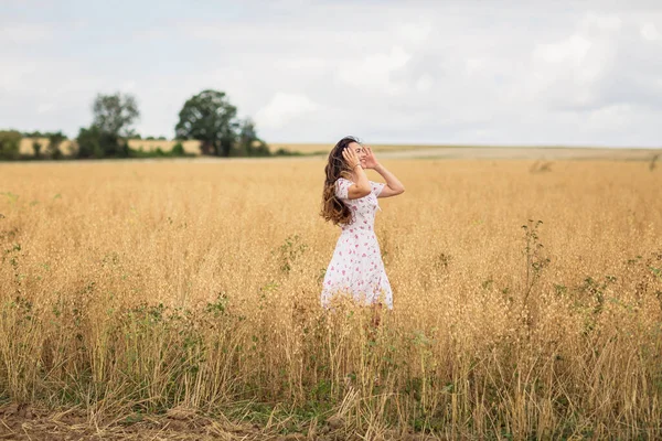 A beautiful brunette woman in a white dress runs along a field of golden chickpeas ear. Stylish girl in the field in windy weather