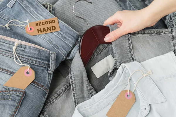 Females clothes on second hand store. Womans hand holding hanger with denim shirt. Craft paper tag with inscription Second hand. Circular fashion, eco friendly shopping, thrift store concept
