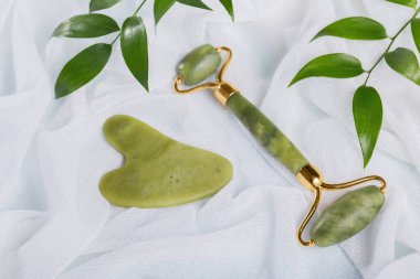 Yeşil yeşim yüzlü silindir ve açık mavi arka planda yaprakları olan gua sha kazıyıcı. Yüz bakımı, spa benliği ve güzellik tedavi konsepti için masaj aracı.