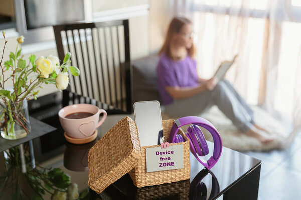 Smartphone is in separate wicker box with inscription Device free zone on table. Woman reading book in background. Stop using digital gadgets. Mental and digital detox concept