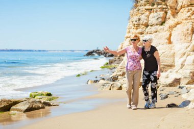 İki yaşlı kadın kayalık sahilde yürüyor, konuşuyor ve gülüyor, birlikte hoş vakit geçiriyorlar.