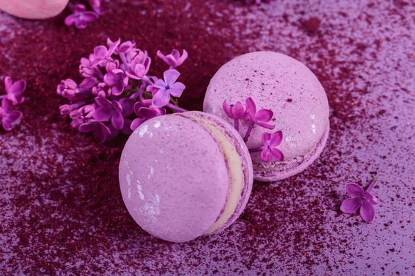 パステルカラーの甘いフランスのマカロンをライラックの花で閉じ 紫色の背景にドライブルーベリーパウダーのスプラッシュ ベーカリーやペストリーショップのための美しい組成 — ストック写真