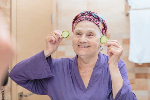 stock image Mature woman taking care of her face. Senior woman standing at the mirror in the bathroom and applying cream or mask on her face and slices of cucumbers on her eyes