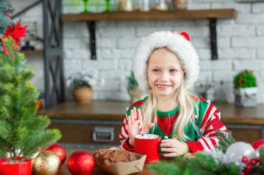 Lifestyle portrait of cute little child girl eating sweet biscuits and drinking warm cacao in mug. Kid sitting in Christmas decorated cozy kitchen and wearing red santa claus hat. 