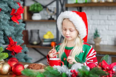 Cute little child girl eating sweet cookies and drinking hot beverages in red cup. Kid sitting in Christmas decorated cozy kitchen and wearing red santa claus hat. New Year winter holidays concept