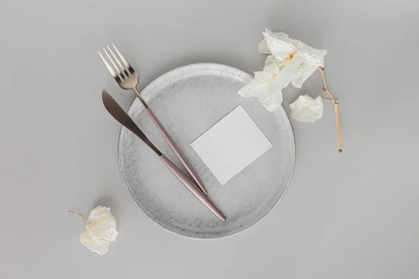 Festive table setting. Golden modern cutlery, dried orchid branch, ceramic plate and blank paper as mock up for invitation or congratulation on white background
