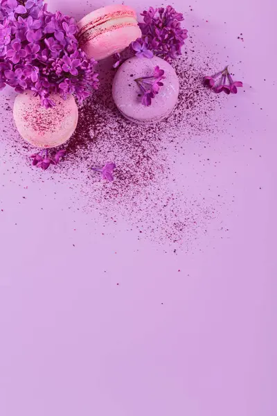 Pastel colored sweet french macaroons with lilac flowers and splash of dry blueberry powder on pink background. Beautiful composition for bakery and pastry shop. Top view with copy space