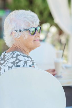 Beautiful senior woman with a cup of coffee with milk resting in nature at the resort in the morning. Vacation, holiday, and travel concept.