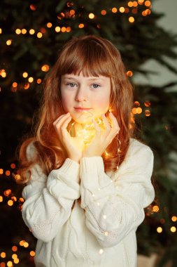 Evin rahat oturma odasında Noel ağacının yanında oturan ve elinde çelenk tutan mutlu genç kız. İyi kış tatilleri.