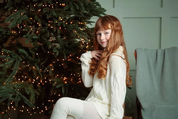 Happy young girl sitting by the Christmas tree in the cozy living room of the house and smiling. Happy winter holidays concept