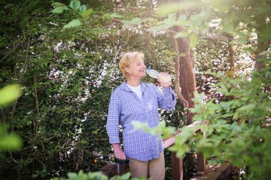 Sarışın, güzel, olgun bir kadın tatillerini dışarıda geçiriyor sırtında sırt çantasıyla ormanda yürüyor, elinde plastik bir şişe su tutuyor ve içiyor. Serinletici, dinlendirici ve aktif yaşlı yaşam tarzı