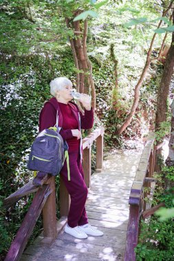 Sarışın, güzel, olgun bir kadın tatillerini dışarıda geçiriyor sırtında sırt çantasıyla ormanda yürüyor, elinde plastik bir şişe su tutuyor ve içiyor. Serinletici, dinlendirici ve aktif yaşlı yaşam tarzı