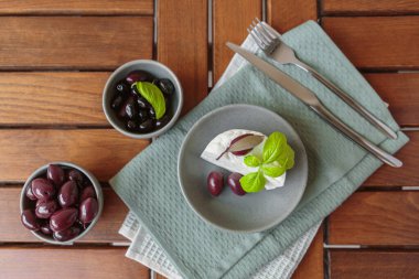 Gri tabakta dilimlenmiş Feta peyniri, siyah zeytin ve fesleğen yapraklarıyla. Terasta lezzetli ve sağlıklı bir kahvaltı.