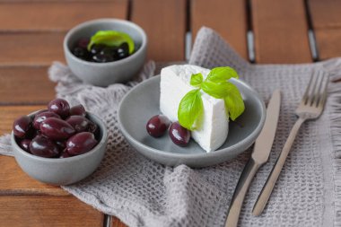 Gri tabakta dilimlenmiş Feta peyniri, siyah zeytin ve fesleğen yapraklarıyla. Terasta lezzetli ve sağlıklı bir kahvaltı.