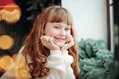 Pencerenin yanında oturan mutlu genç kız ve evin rahat oturma odasında Noel ağacı. İyi kış tatilleri.