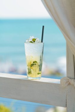 Mavi denize karşı terasta bir bardak Majito kokteyli. Sıcak güneşli bir günde, deniz kenarındaki kafede soğuk bir içecek.