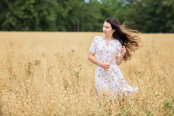 Young woman in white midi dress walking across the field with ripe golden ears of chickpeas. Beauty Romantic woman Outdoors