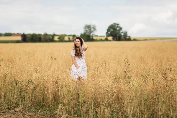 A beautiful brunette woman in a white dress runs along a field of golden chickpeas ear. Stylish girl in the field in windy weather