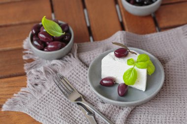 Gri tabakta dilimlenmiş Feta peyniri, siyah zeytin ve fesleğen yapraklarıyla. Terasta lezzetli ve sağlıklı bir kahvaltı.
