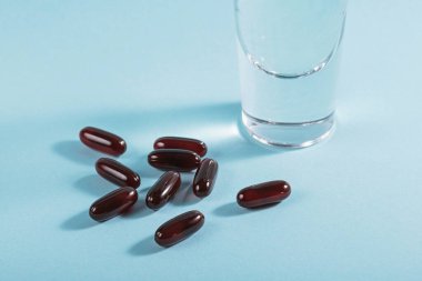 Diyet, beslenme, sağlıklı yeme konsepti. Açık mavi zemin üzerine kahverengi softgel kapsülleri dökmek için gıda takviyeleri ve bir bardak su dolu yağlı kapsüller..