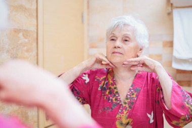 Mature woman taking care of her face. Senior woman standing at the mirror in the bathroom and applying cream to her face or doing self-massage