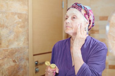 Yüzüne bakan yaşlı bir kadın. Banyoda aynanın karşısında duran ve yüzüne krem ya da maske süren son sınıf öğrencisi kadın.