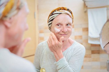 Olgun bir kadın yüzüne bakıyor. Banyoda aynanın karşısında duran ve yüzüne krem ya da maske süren son sınıf öğrencisi kadın.