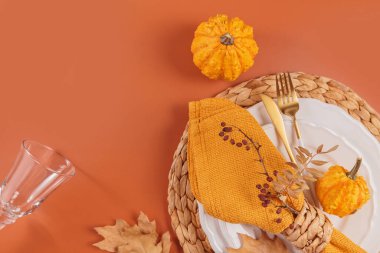 Turuncu arka planda balkabağı süslemeleri olan sonbahar masası. Boş tabak, bardak, çatal bıçak takımı, sarı keten peçete ve kuru yapraklar. Sonbahar tatili, Cadılar Bayramı, Şükran Günü konsepti