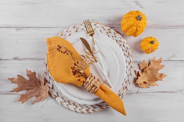 Autumn table setting with pumpkin decorations on white wooden background. Empty plate, cutlery, yellow linen napkin and dry leaves. Autumn holiday mood, Halloween, Thanksgiving concept. Top view