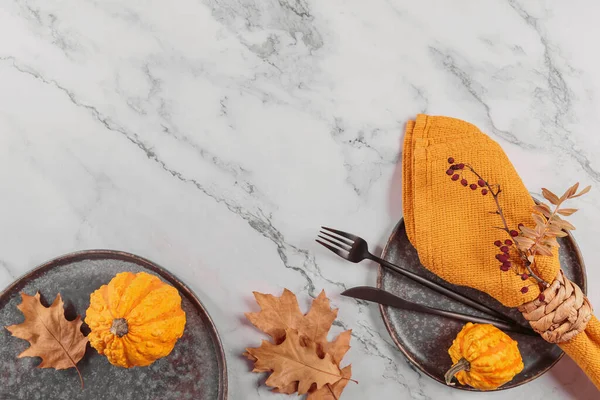 Autumn table setting with pumpkin decorations on light background. Dark plate, cutlery, yellow linen napkin and dry leaves. Autumn holiday mood, Halloween, Thanksgiving concept. Top view, flat lay