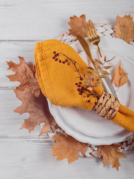 Autumn table setting with decorations on white wooden background. Empty plate, cutlery, yellow linen napkin and dry leaves. Autumn holiday mood, Halloween, Thanksgiving concept. Top view