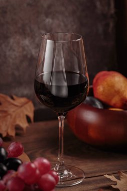 Koyu ahşap arka planda meyvelerle servis edilen bir bardak zengin kırmızı şarap. Sonbahar, şarap, üzüm ve kuru yapraklarla dolu kırsal bir hayat. Süveter hava konsepti
