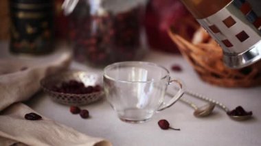 Etrafında yabani sonbahar meyveleri olan şeffaf cam gül kalçası çayı. Çaydanlıktan sıcak içecek dökülüyor. Sıcak kış içeceği