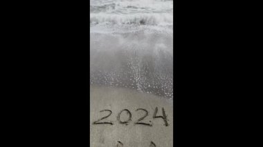 Takvim ve Yeni Yıl 2024 konsept videosu. Metin 2024 koyu kum yüzeyinde elle yazılmış. Deniz dalgası plajda rakamları silip süpürüyor.