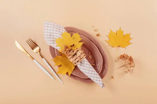 Autumn table setting on beige background. Oval plates, cutlery, checkered napkin and dry yellow leaves. Autumn holiday mood, Halloween, Thanksgiving concept. Top view