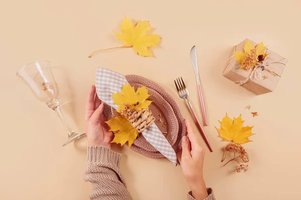 Autumn table setting on beige background. Womens hands holding oval plates, cutlery, checkered napkin, gift box and dry yellow leaves. Autumn holiday mood, Halloween, Thanksgiving concept. Top view
