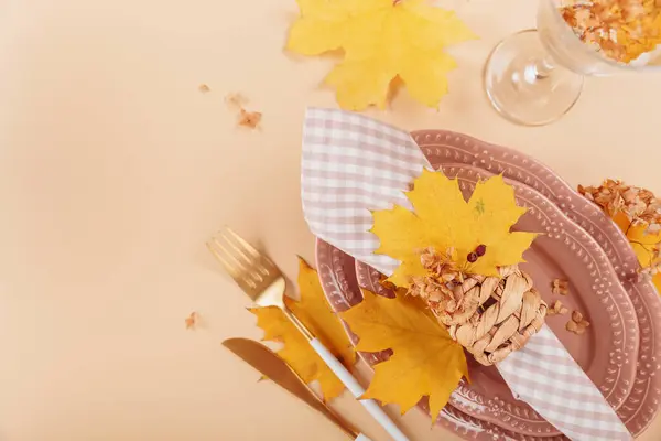 Autumn table setting on beige background. Oval plates, cutlery, glass, checkered napkin and dry yellow leaves. Autumn holiday mood, Halloween, Thanksgiving concept. Top view, copy space