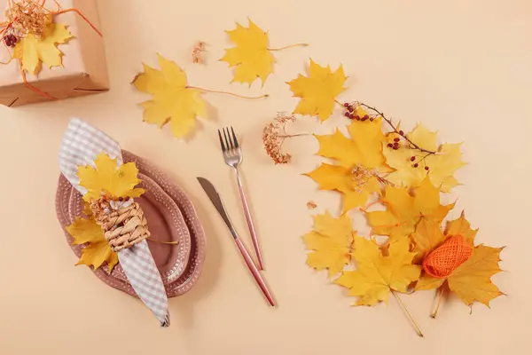 Autumn table setting on beige background. Oval plates, cutlery, checkered napkin, gift box and dry yellow leaves. Autumn holiday mood, Halloween, Thanksgiving concept. Top view, flat lay