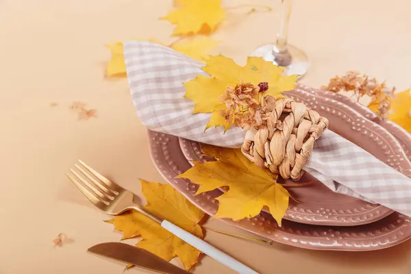 Autumn table setting on beige background. Oval plates, cutlery, glass, checkered napkin and dry yellow leaves. Autumn holiday mood, Halloween, Thanksgiving concept. Close up