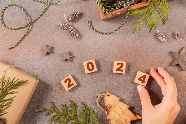 Winter holiday mood, Merry Christmas, Happy New Year background. Preparation for holidays. Handmade gift box, wooden box with Christmas decor and womans hand adding up numbers 2024. Flat lay, top view